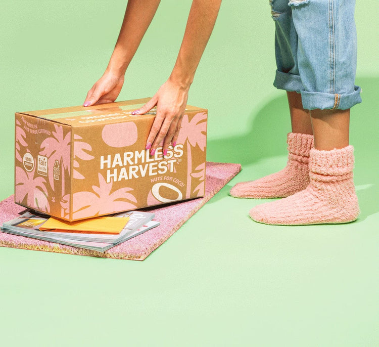 Person with pink socks reaching for box of Harmless Harvest Organic Coconut Water on doormat